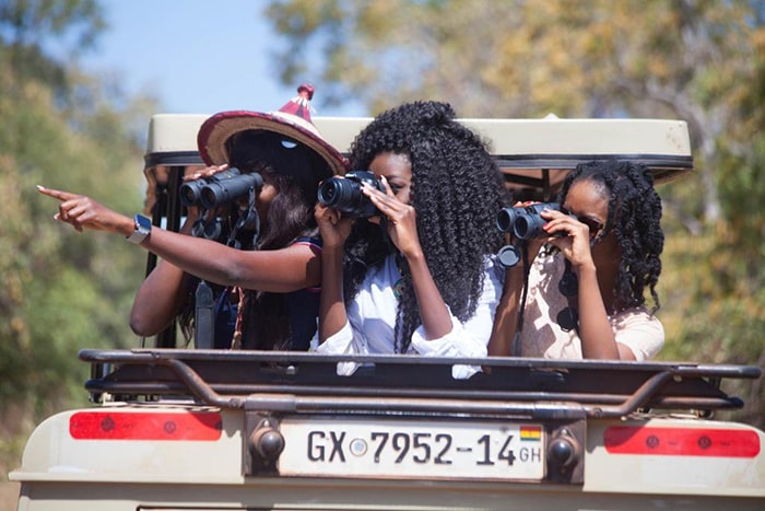 Image of three young beautiful African ladies standing at the back of a Zaina Lodge game drive vehicle, looking through binoculars to spot animals in Mole National Park